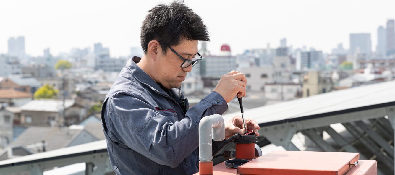 屋上の設備を真剣な表情で点検・メンテナンスする、作業服姿の技術職の男性。