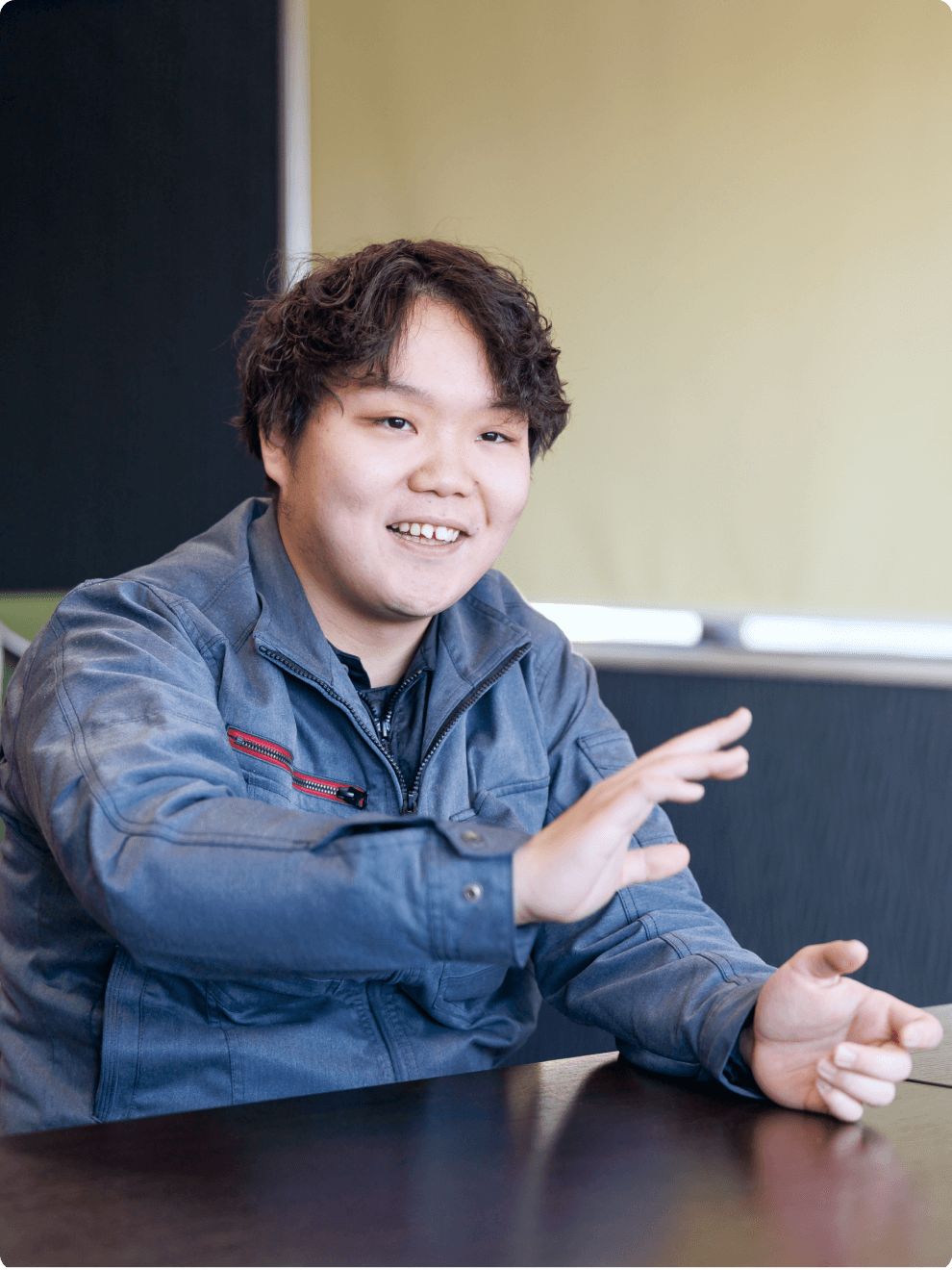 笑顔で身振り手振りを交えながらインタビューに答える、グレーの作業服を着た若手男性社員。