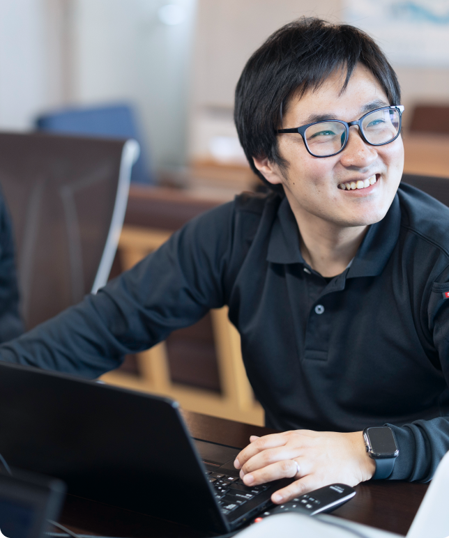 ノートパソコンを前に笑顔で業務に取り組む、眼鏡をかけた男性社員。風通しの良いオフィスでの日常的な仕事風景。