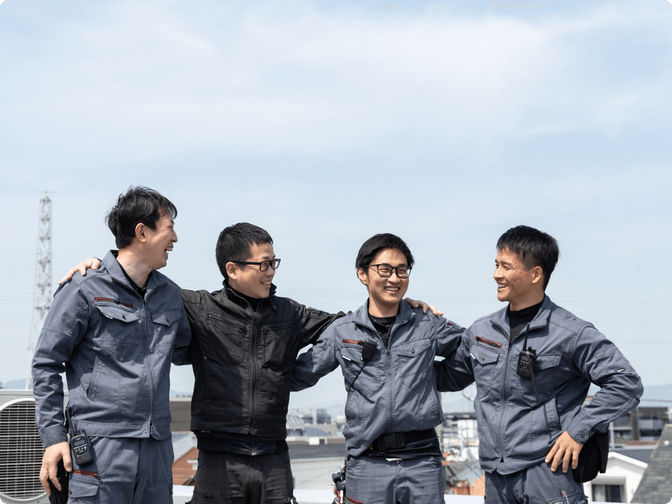 青空の下、屋上で肩を組みながら笑顔で談笑する、チームワーク抜群な4人の作業スタッフたち。