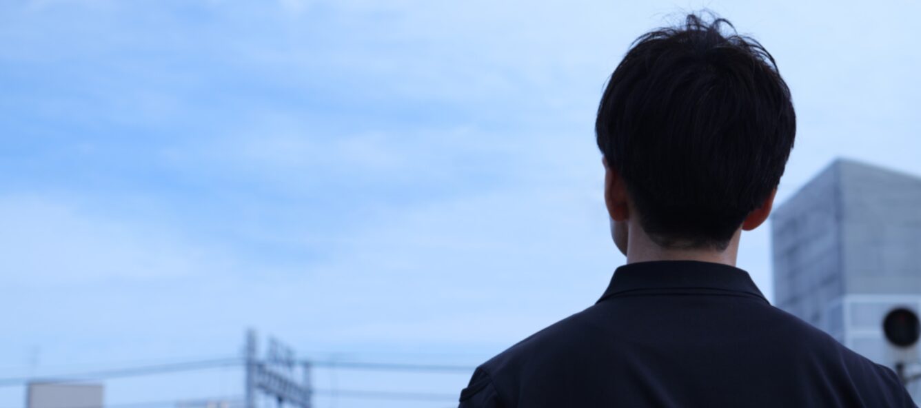 青空を背景に、将来を見据えるように街並みを眺めて佇む男性スタッフの背中。