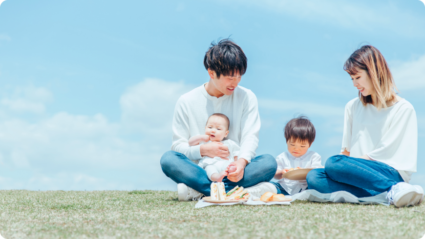 澄み渡る青空の下、芝生でピクニックを楽しむ笑顔の4人家族。仕事とプライベートの両立を大切にする、温かな職場文化。