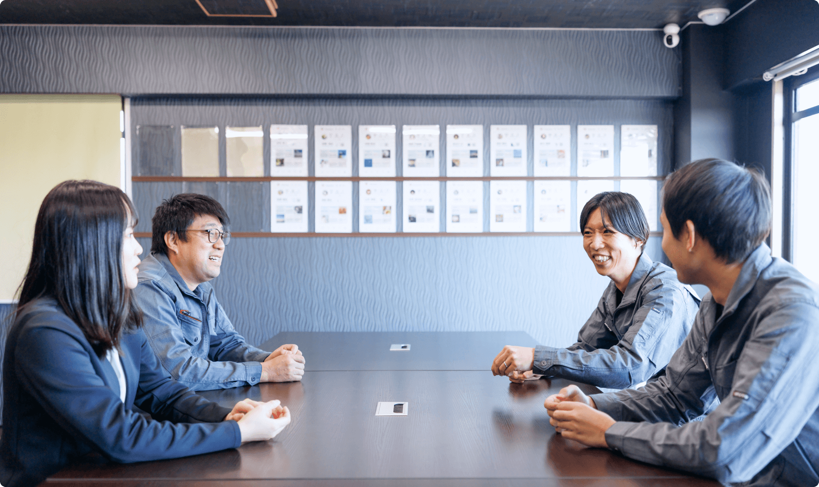 オフィスでの打ち合わせ中、笑顔で意見を交わす4名の社員。部署の垣根を超えたコミュニケーションと風通しの良い職場環境。