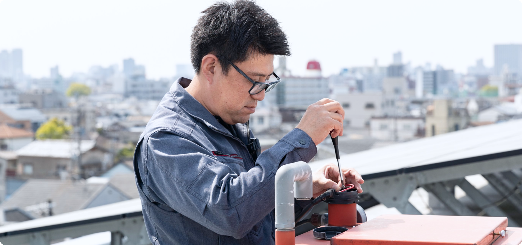屋上の設備を工具で精密にメンテナンスする技術者。都市のインフラを支える「建物の守護者」としての責任感ある作業風景。