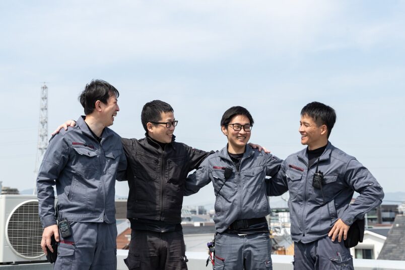 青空の下、屋上で肩を組みながら笑顔で談笑する、チームワーク抜群な4人の作業スタッフたち。