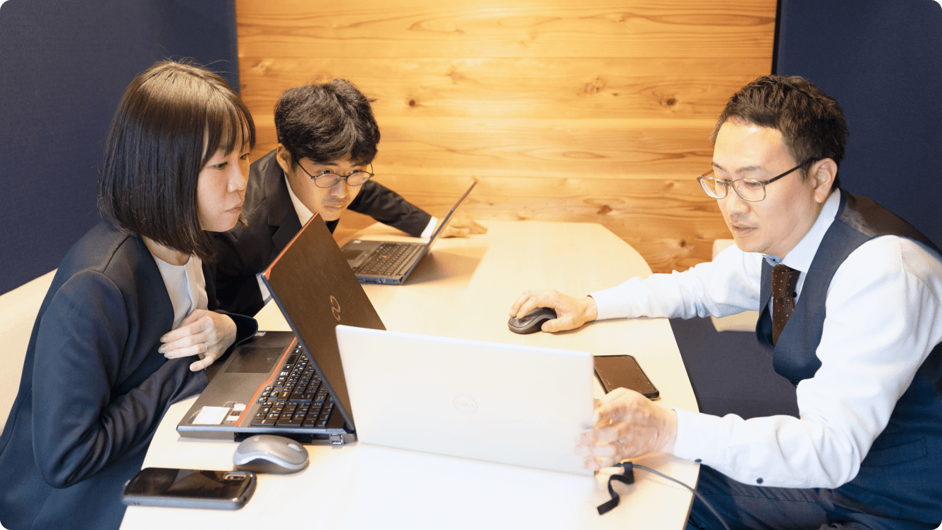 木目調のデスクでPCを使い、真剣な表情で技術的な詳細を詰め合う社員たち。確実な施工を実現するための正確なチームプレイ。