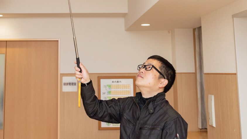 点検棒を使用して天井の火災報知器を慎重に検査する点検員。見えないリスクを未然に防ぐ、徹底した保守管理。
