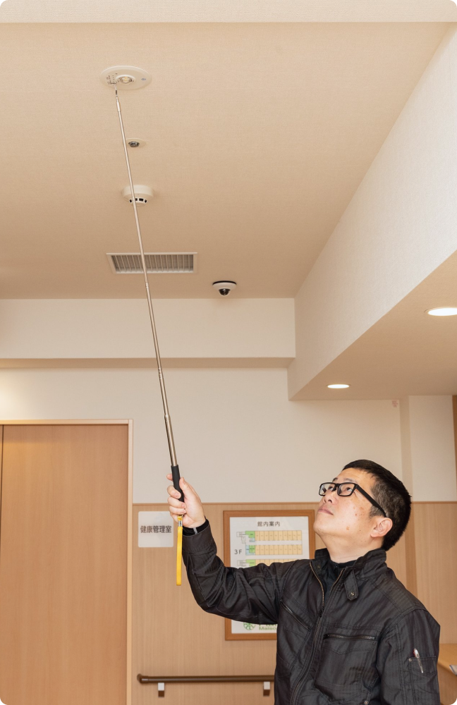 点検棒を使用して天井の火災報知器を慎重に検査する点検員。見えないリスクを未然に防ぐ、徹底した保守管理。