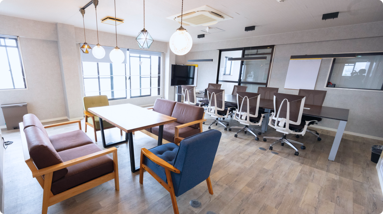 木目調のフロアにモダンなペンダントライトが下がる、カフェのような雰囲気のミーティングスペース。手前にはソファ席、奥にはデスクとオフィスチェアが並ぶ。