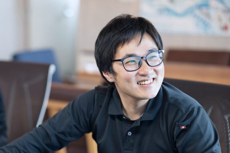 ノートパソコンを前に笑顔で業務に取り組む、眼鏡をかけた男性社員。風通しの良いオフィスでの日常的な仕事風景。